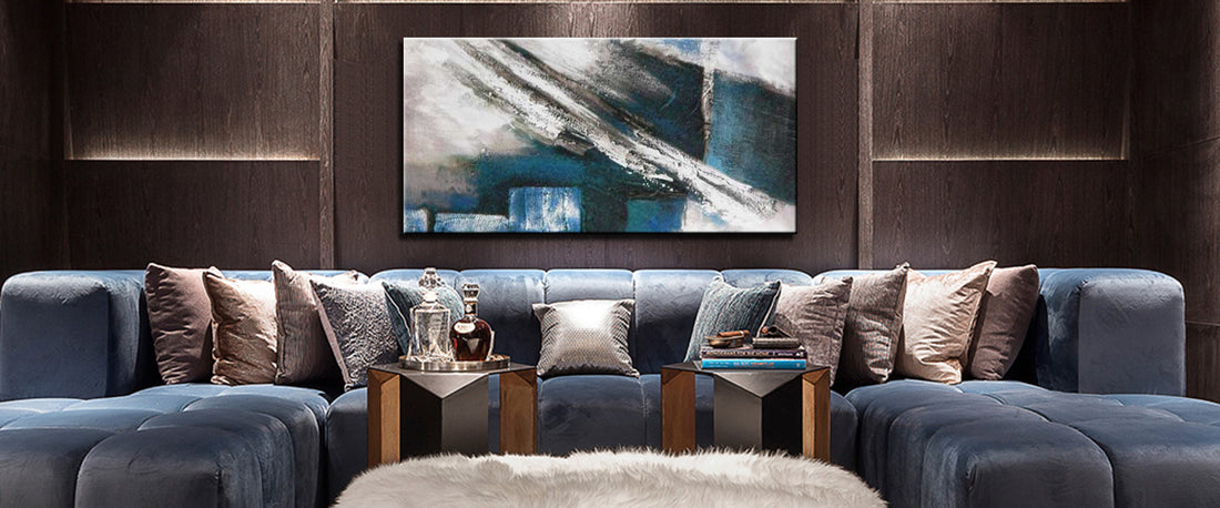 Diepblauwe U-vormige bank met neutrale kussens, geometrische bijzettafels met glaswerk en boeken, en abstract schilderij in blauw en houttinten erboven.